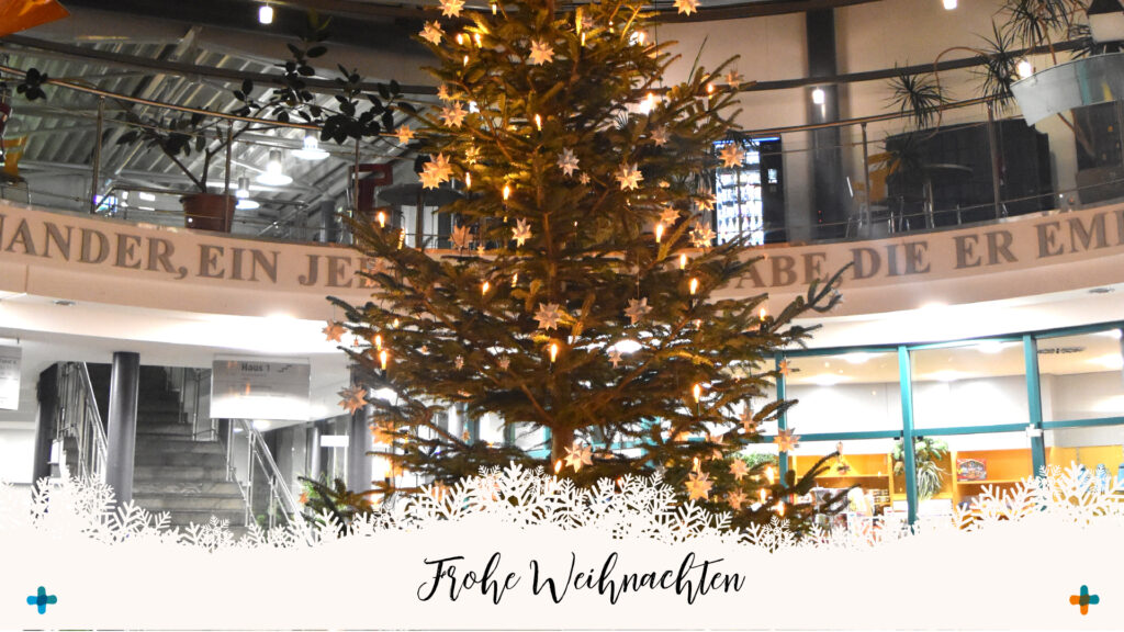 Ein festlich geschmückter Weihnachtsbaum mit warmen Lichtern und Sternen in einem modernen Foyer (Klinikum). Im unteren Bereich befindet sich eine weiße Schneeflocken-Grafik mit dem geschwungenen Schriftzug „Frohe Weihnachten“