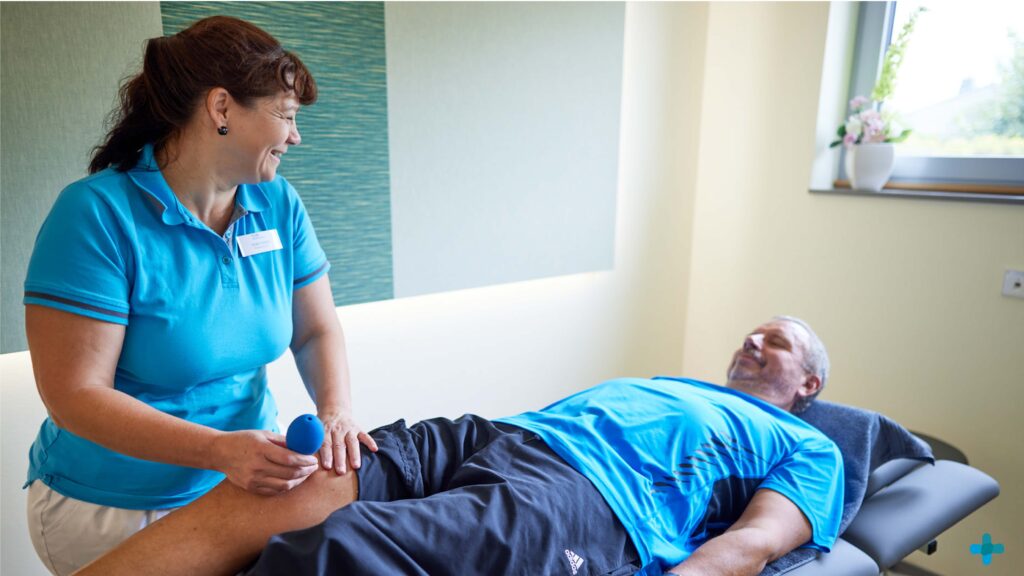 Eine Physiotherapeutin behandelt die Oberschenkelmuskulatur eines Patienten auf einer Therapieliege mit einem Massageball.