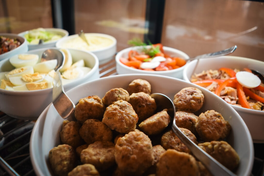 Auswahl an warmen und kalten Speisen in einer Vitrine, darunter eine große Schale mit Frikadellen, gekochte Eier und Tomaten-Mozzarella-Salat.