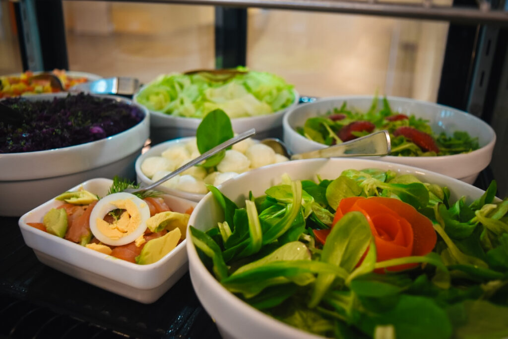 Auswahl an frischen Salaten wie Feldsalat mit Tomatenrose, Mozzarellabällchen und Ei-Avocado-Salat in weißen Schalen in einer Vitrine.