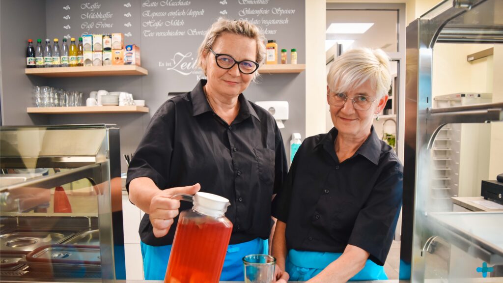 Zwei lächelnde Mitarbeiterinnen in schwarzer Arbeitskleidung und blauen Schürzen stehen hinter einer Theke; eine gießt aus einer Glaskaraffe ein rötliches Kaltgetränk in ein Glas.
