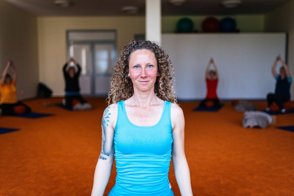 Yogalehrerin lächelt im Vordergrund eines Kursraums, im Hintergrund machen Teilnehmer Yogaübungen auf Matten