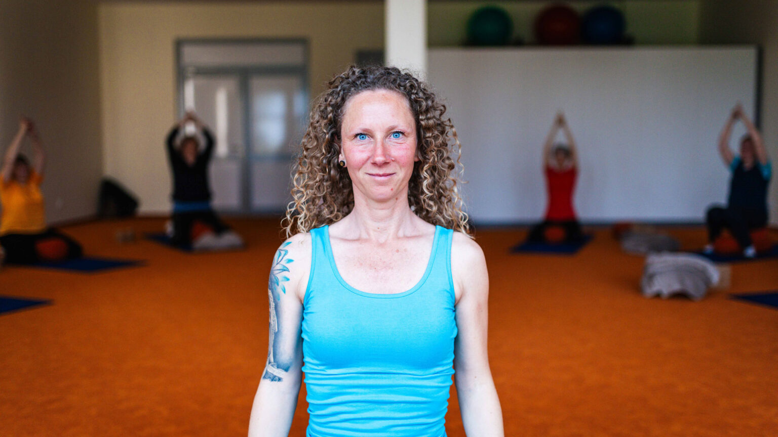 Yogalehrerin lächelt im Vordergrund eines Kursraums, im Hintergrund machen Teilnehmer Yogaübungen auf Matten